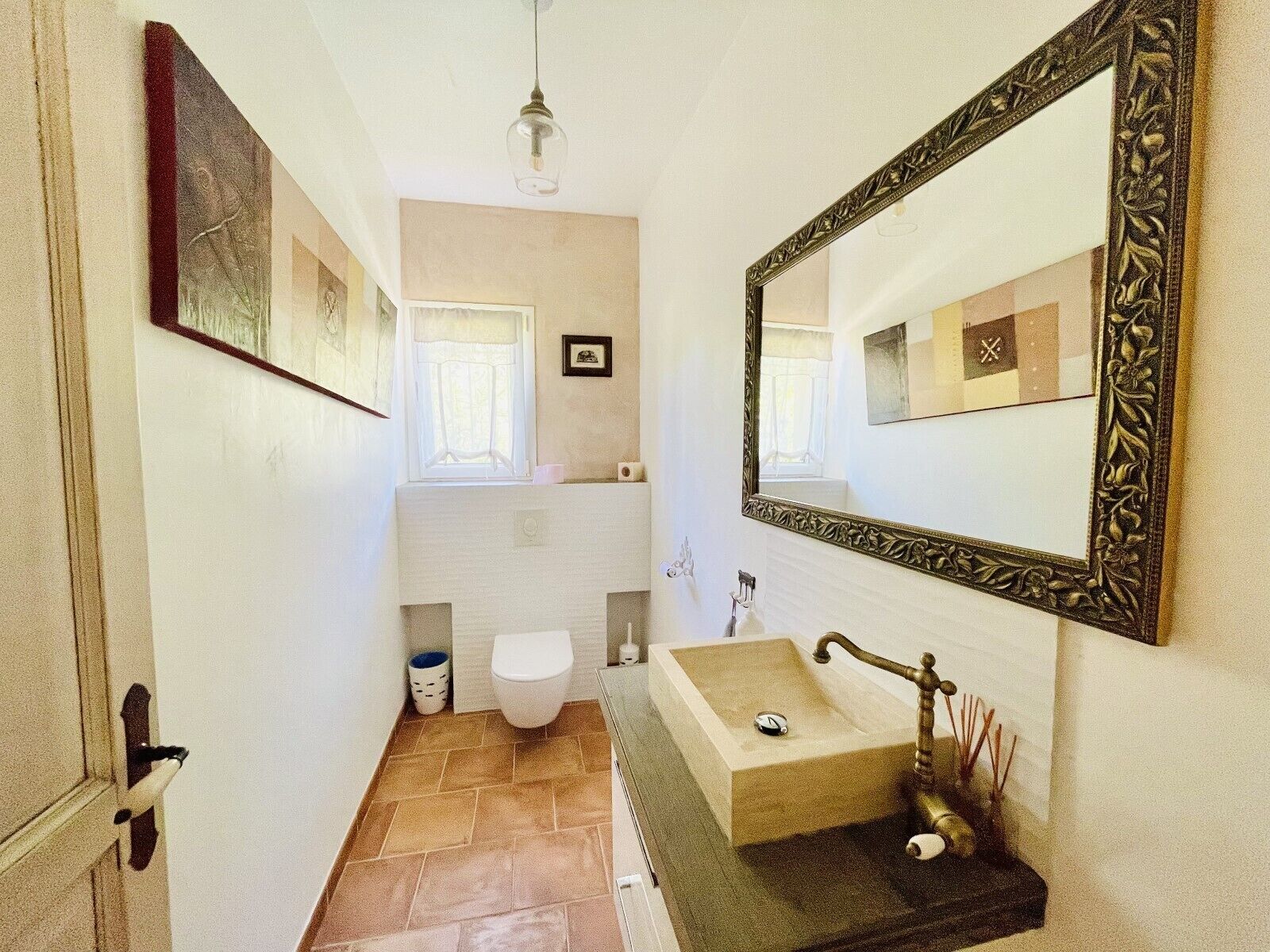 A narrow bathroom with a white toilet, a beige sink with a brass faucet, and a large mirror with an ornate frame.