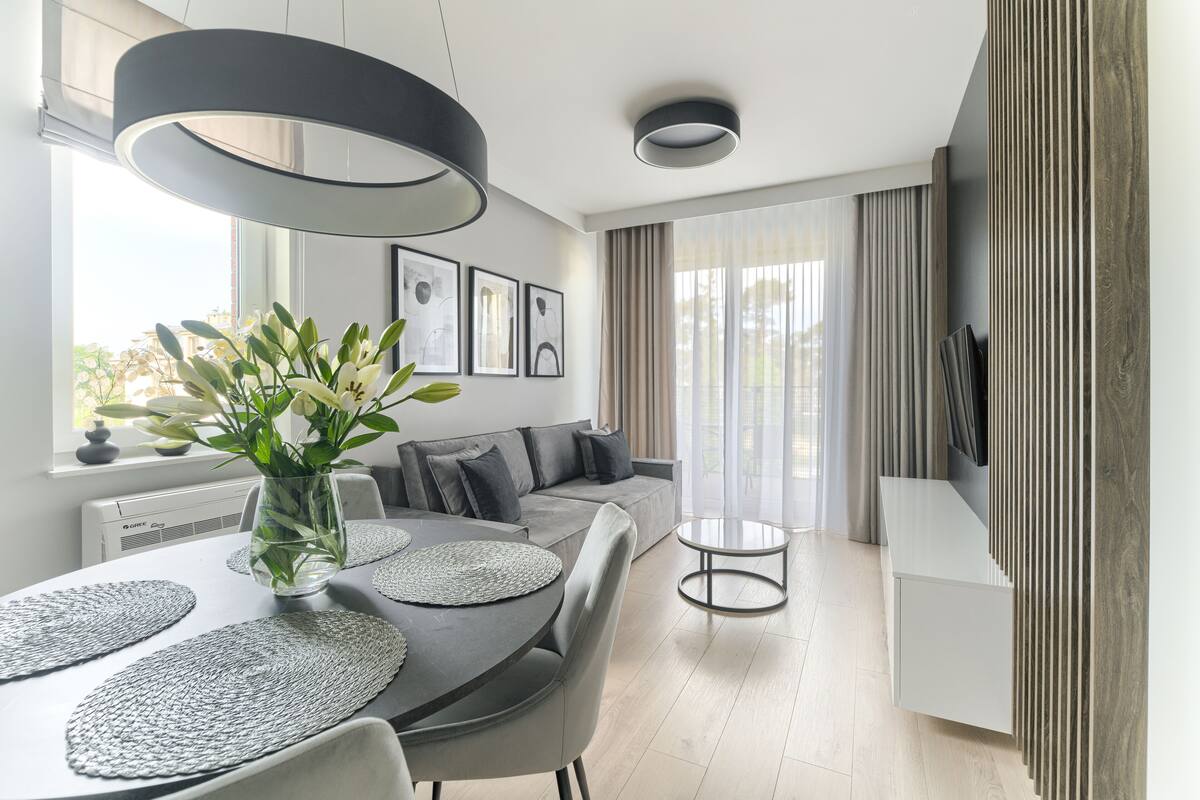 A modern living room with a grey sofa, a dining table with placemats, a vase with flowers, and a TV on the wall.