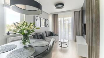 A modern living room with a grey sofa, a dining table with placemats, a vase with flowers, and a TV on the wall.