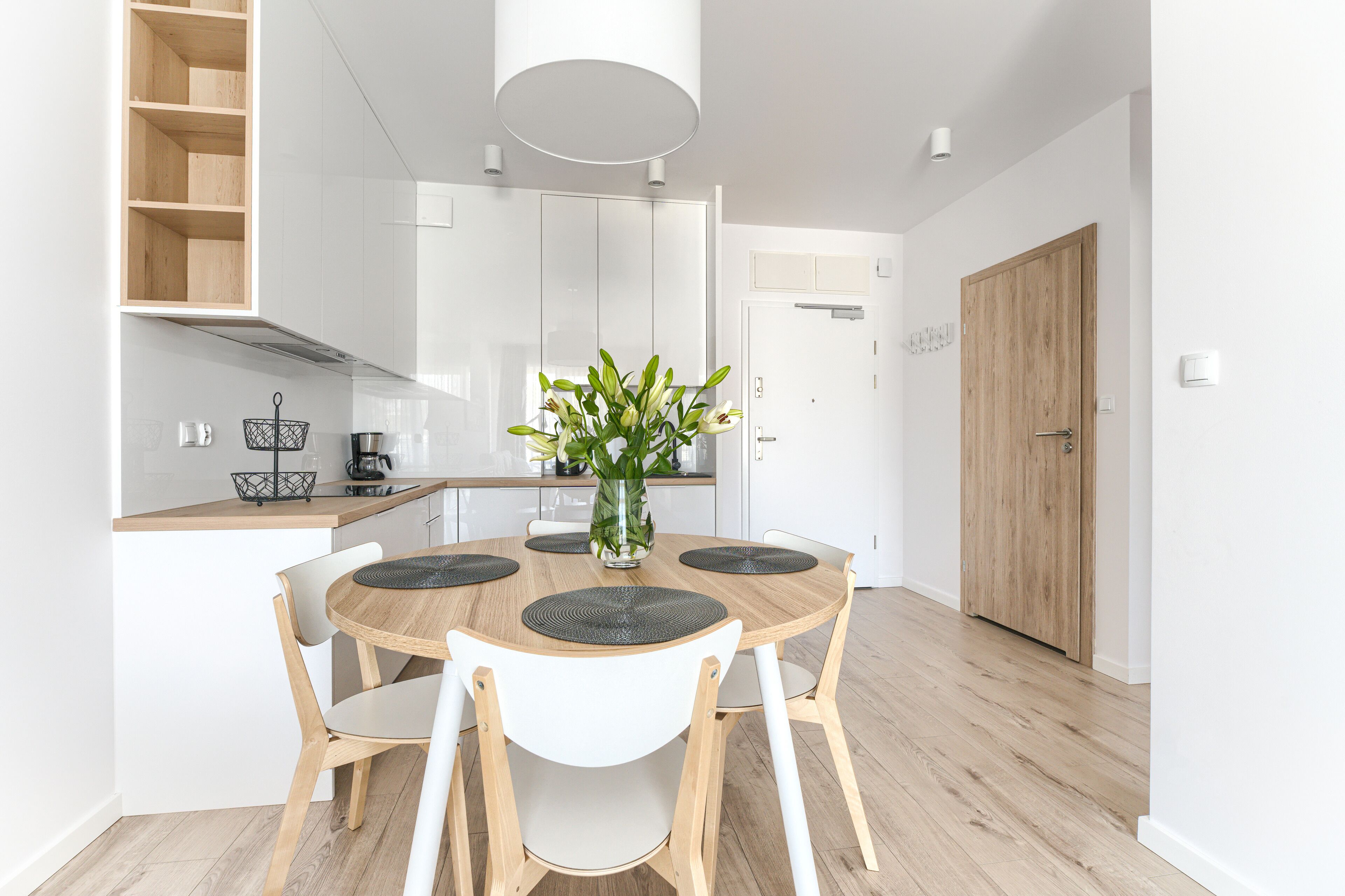 A modern kitchen with white cabinetry, a wooden dining table with four chairs, a vase with flowers, and a wooden door.