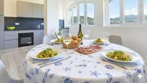 Un comedor con una mesa redonda dispuesta para cuatro personas, con platos de pasta, pan y una botella de vino. La habitación tiene ventanas amplias con vistas a colinas y una cocina equipada con electrodomésticos modernos.