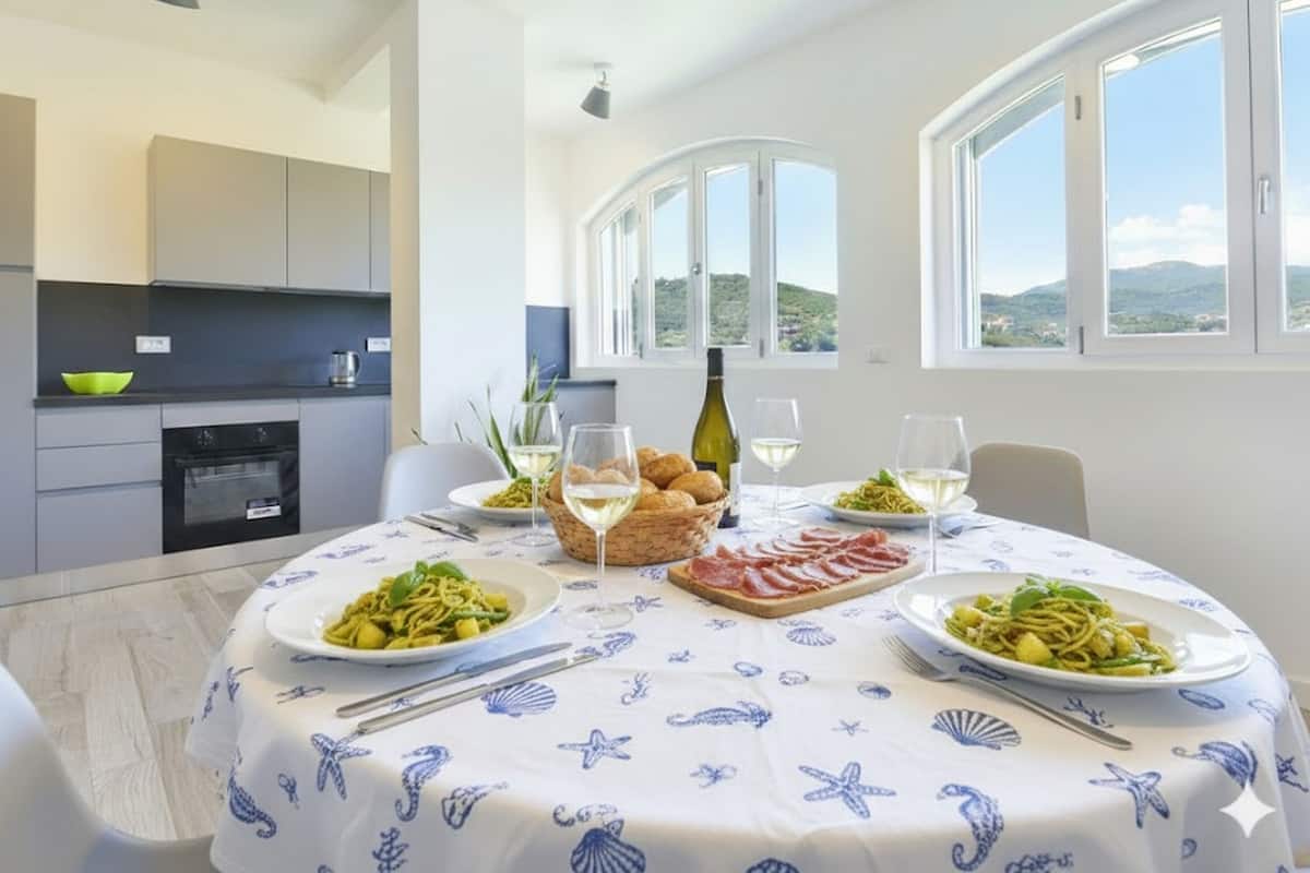 A dining area with a round table set for four, featuring plates of pasta, bread, and a bottle of wine. The room has large windows with a view of hills and a kitchen area with modern appliances.