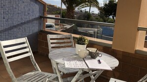 Une terrasse avec des chaises et une table en métal blanc, un pot de fleurs et, en arrière-plan, une vue sur des palmiers et un bâtiment.