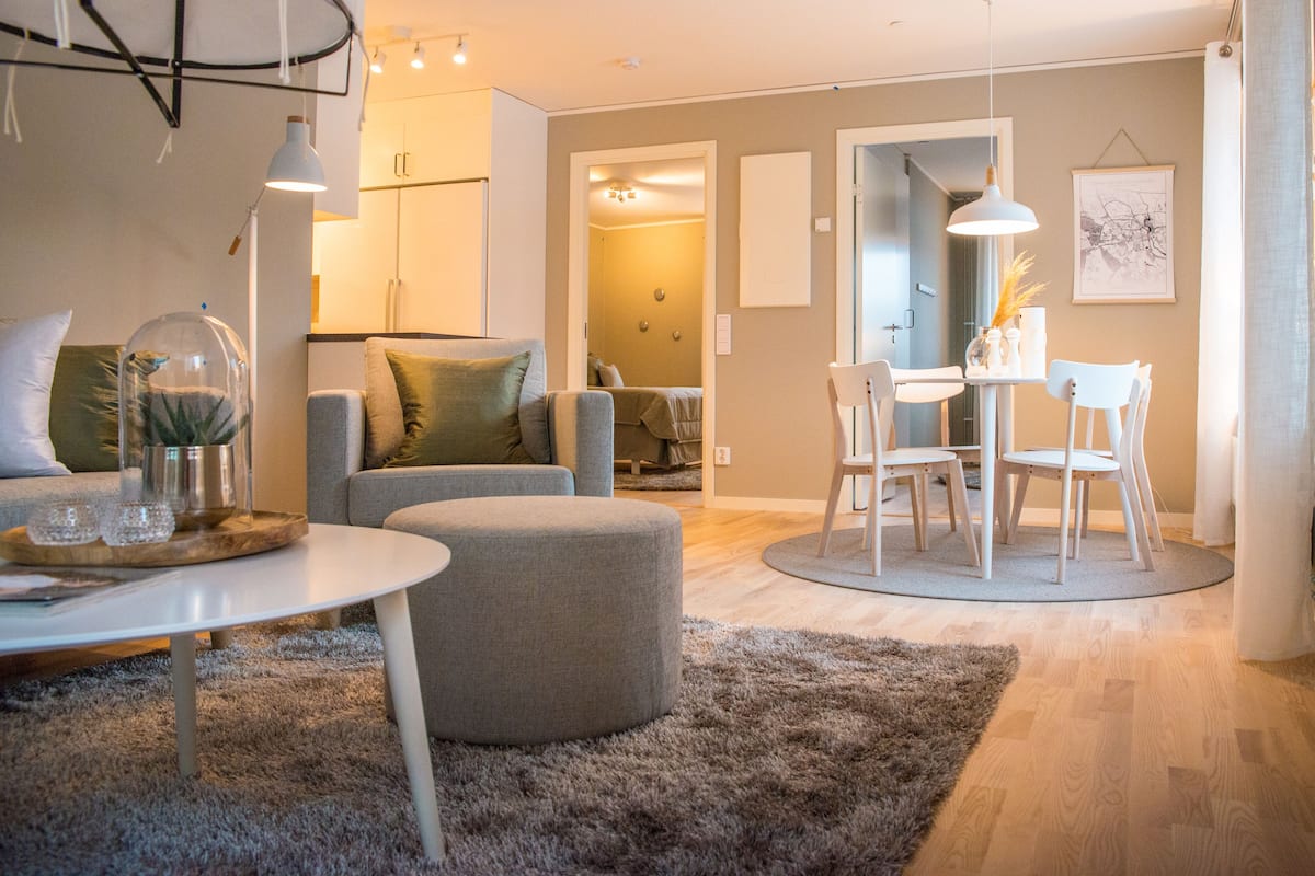 Uma sala de estar moderna com uma mesa de centro redonda, sofá cinza e um banquinho. Há uma área de jantar com uma mesa e cadeiras brancas.