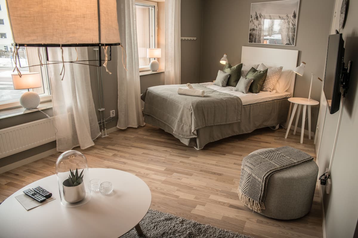 Quarto moderno com cama, luminárias de cabeceira, TV e uma mesinha com uma planta e controles remotos.