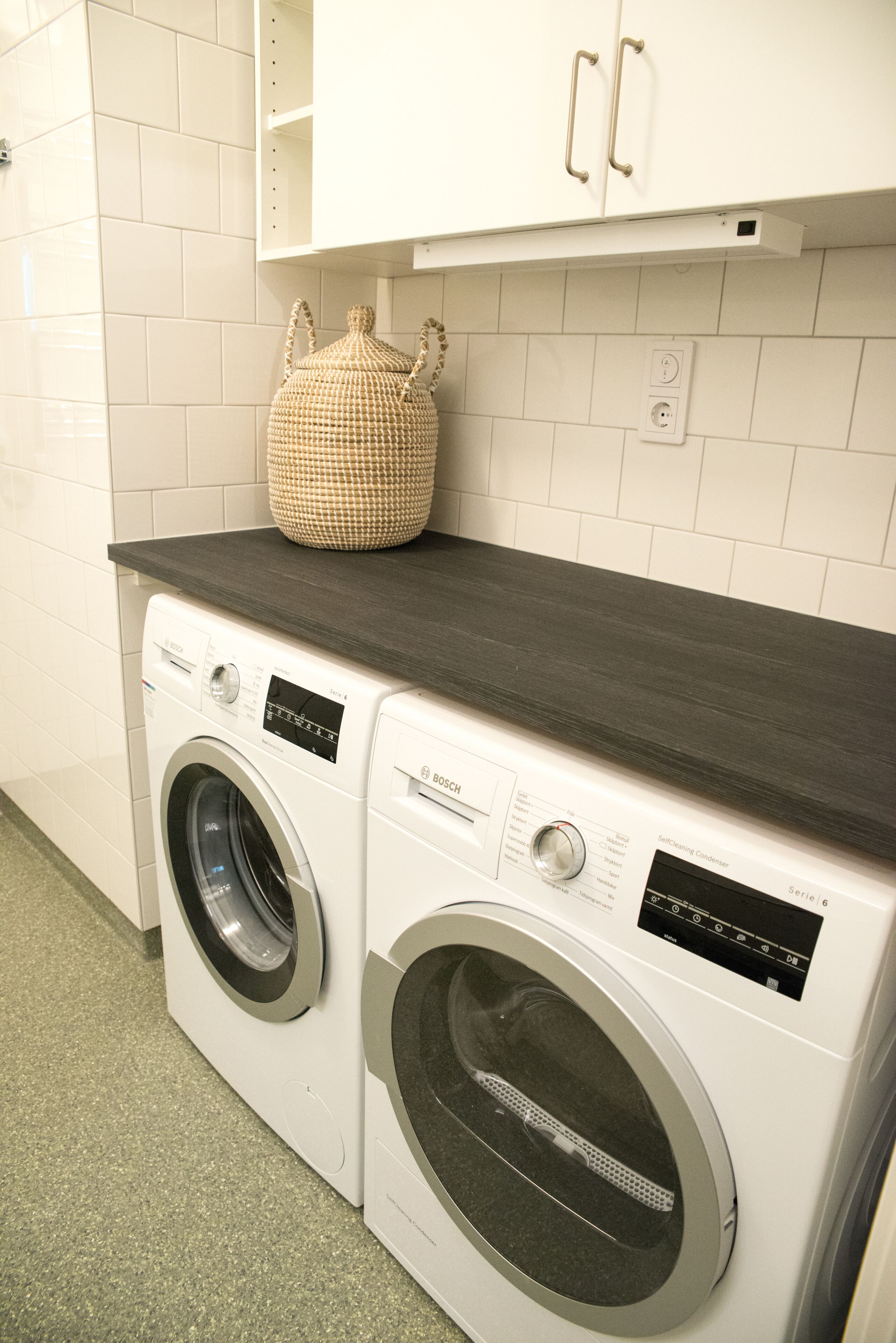 A laundry area with two front-loading washing machines, a dark countertop, and a white tiled wall.