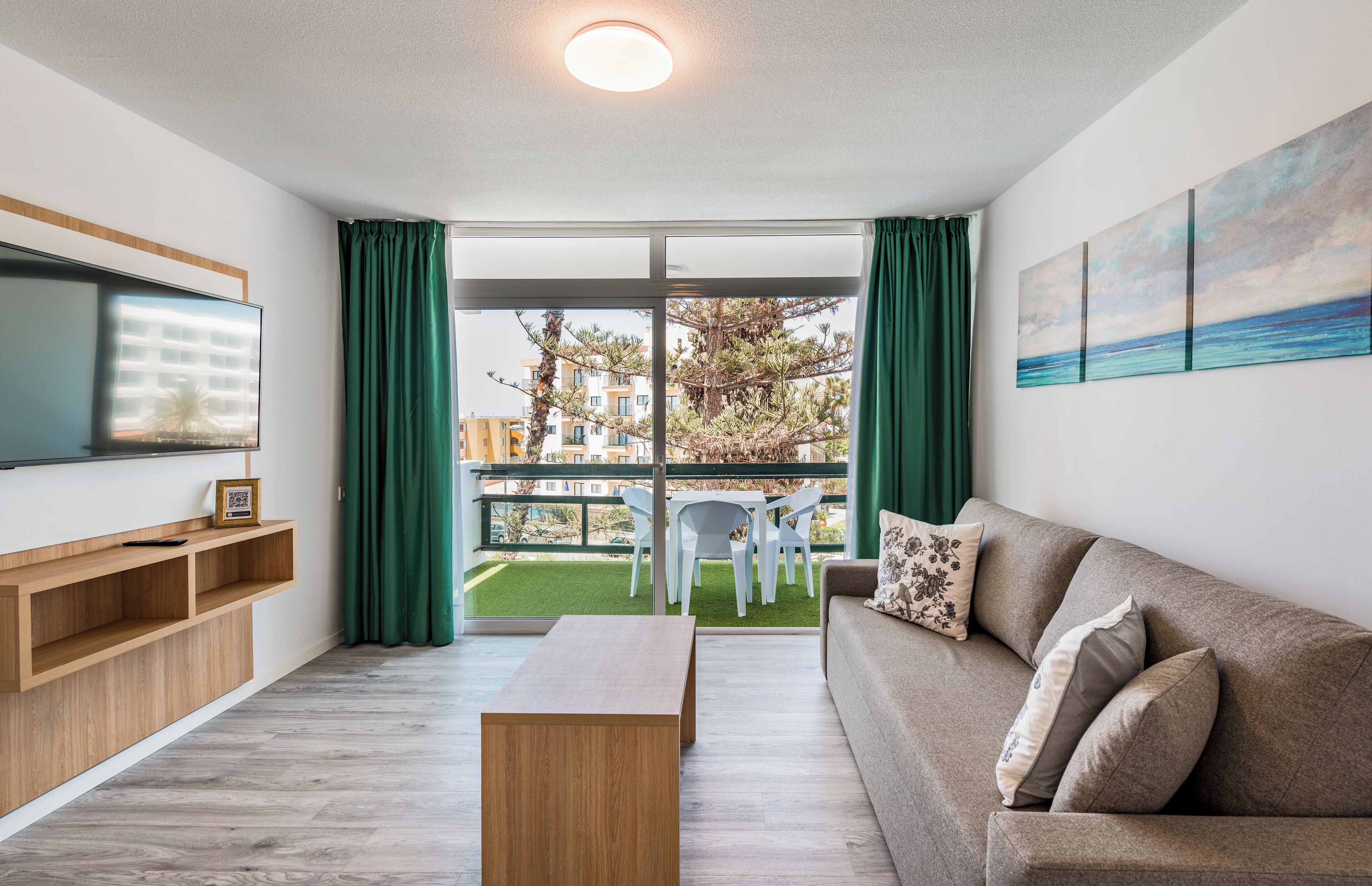 Una sala de estar moderna con un sofá, una mesa de centro, un televisor y un balcón con vistas a edificios y árboles.