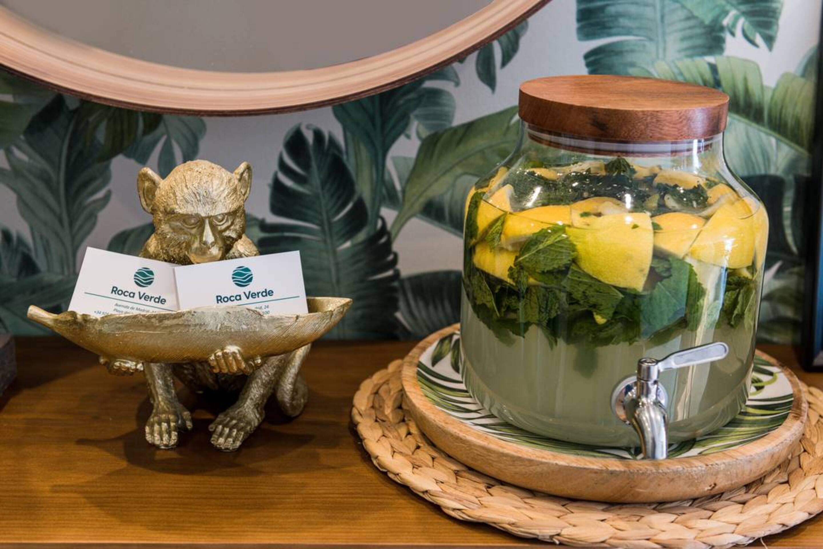 Een decoratief tafereel met een glazen pot gevuld met geel fruit en groene bladeren, een gouden beeldje dat een kaart vasthoudt, en een houten kom op een geweven onderzetter.