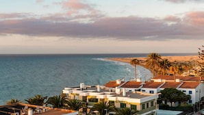 Vista costera con un edificio de techo plano y antenas satelitales, con vista al mar y a una playa de arena con palmeras.