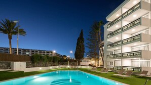 Una piscina con sillas de descanso y un edificio de varios pisos con balcones.
