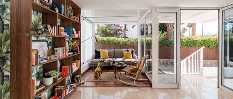 A modern living room with a wooden bookshelf, a sofa with yellow cushions, a rocking chair, and a glass door leading to a balcony with greenery.