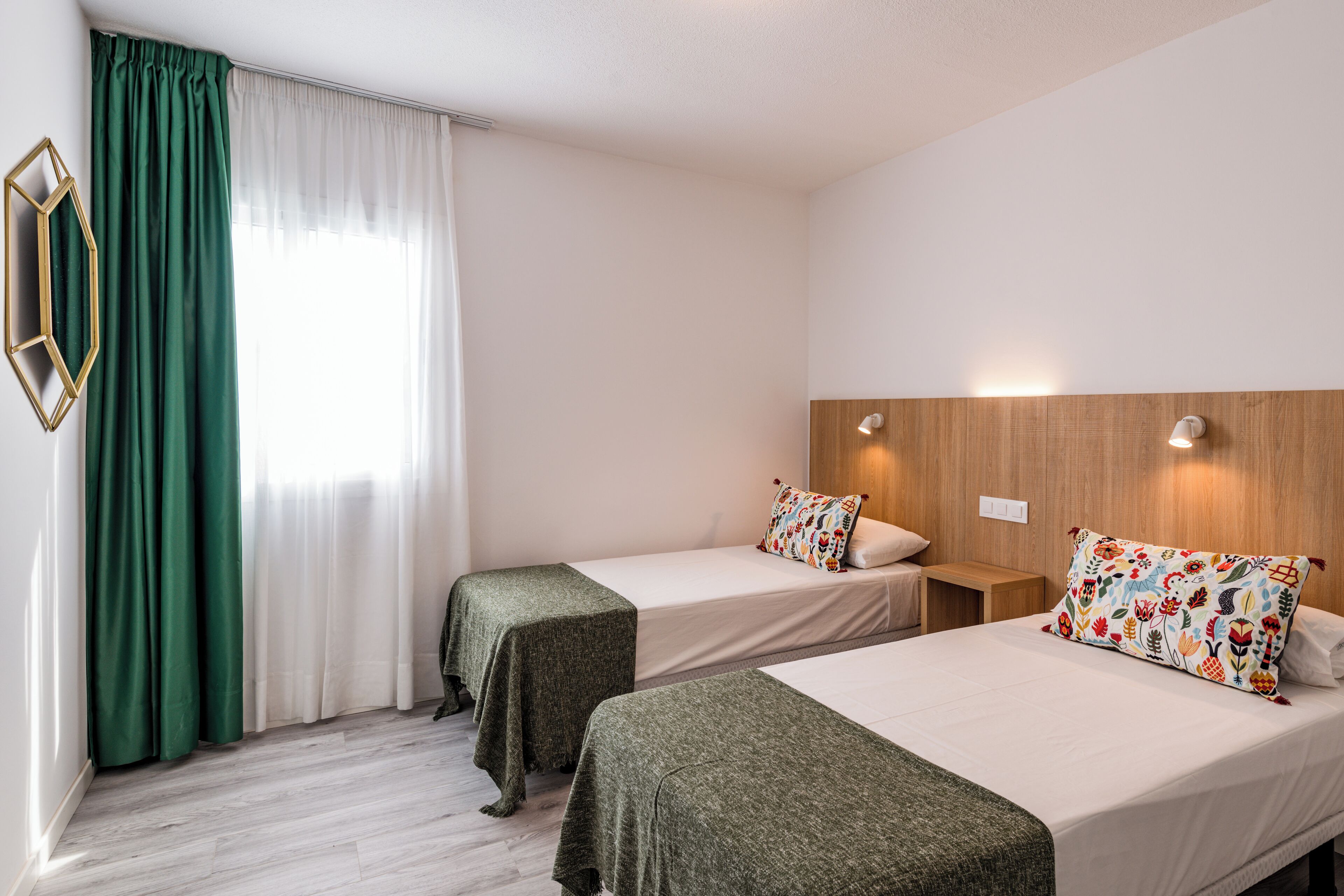 Hotellihuone, jossa on kaksi erillistä sänkyä, puinen pääty, peili ja ikkuna, jossa on vihreät verhot.