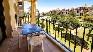 A balcony with a table and chairs, overlooking a well-maintained garden and a row of buildings. - Alcaudon - Isla Canela Beach IAHL59P273 (Isla Canela)