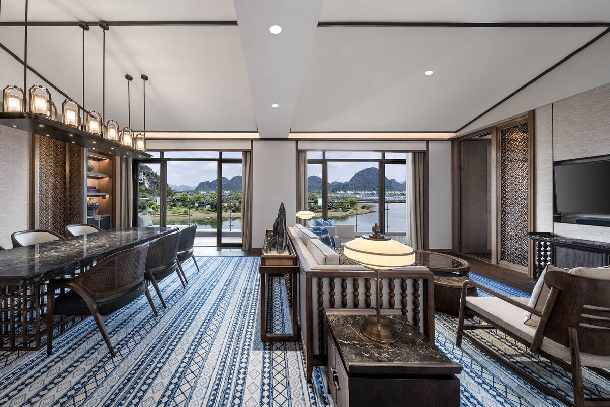 A modern hotel room with a dining area, a sofa, a TV, and a view of a mountainous landscape.