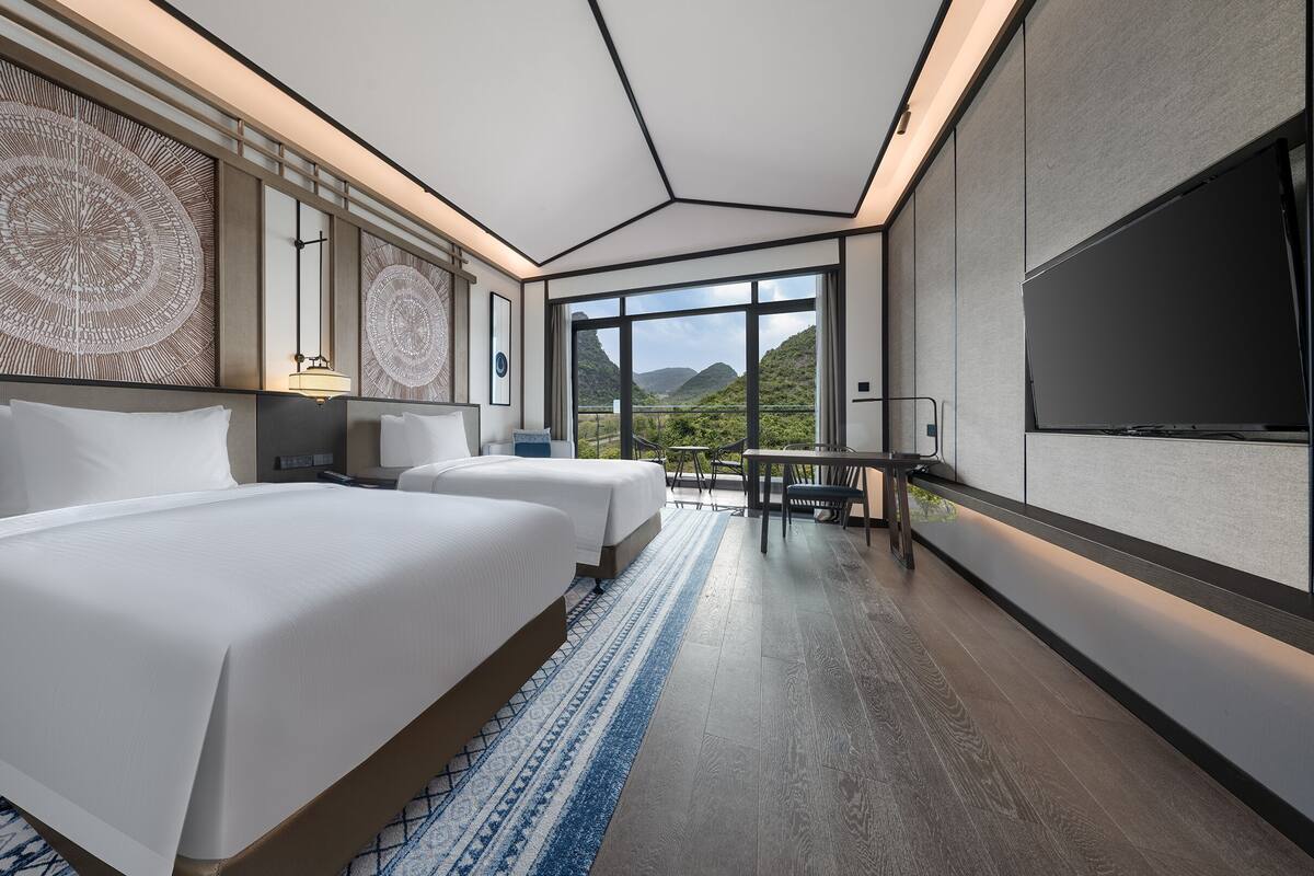 A modern hotel room with two beds, a TV, and a balcony with a mountain view.
