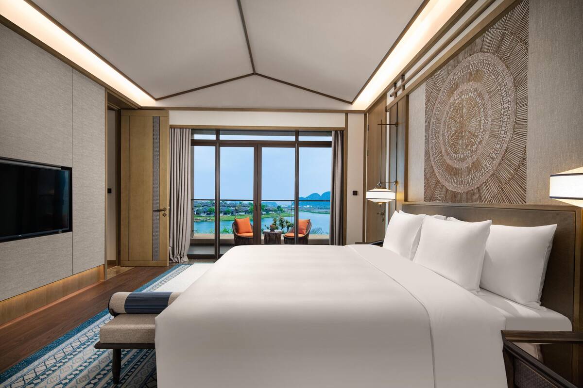 A hotel room with a large bed, a TV, a view of a body of water, and a balcony with chairs.