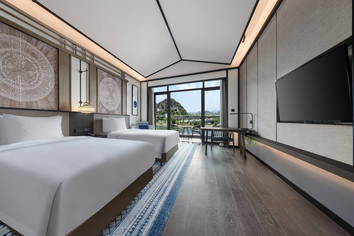 A modern hotel room with two beds, a large TV, and a view of a mountainous landscape.