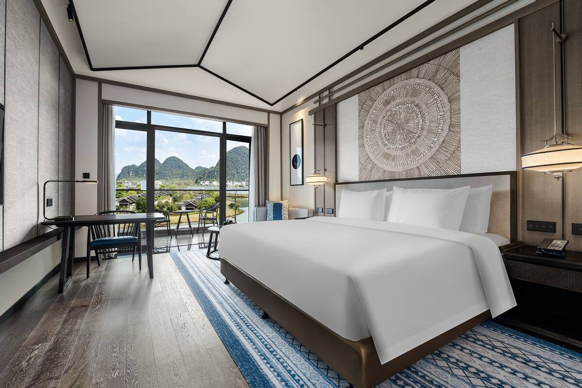 A modern hotel room with a large bed, a desk, and a view of a mountainous landscape.