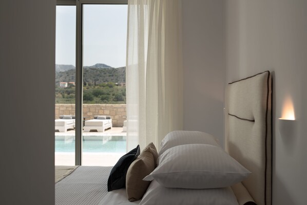 Un dormitorio con cama, almohadas, cabecera y vistas a una piscina y montañas.