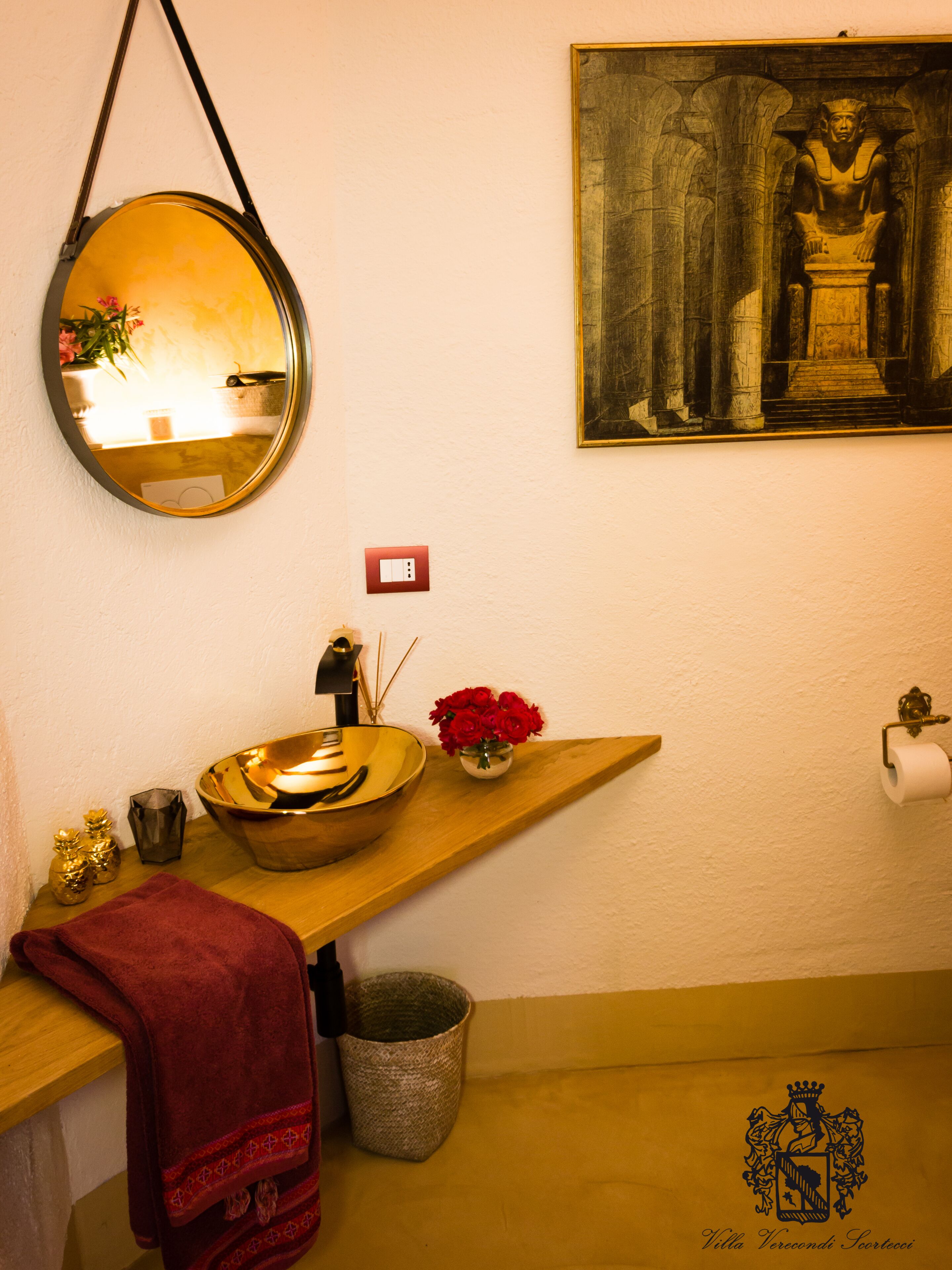 A bathroom with a round mirror, a sink with a bowl, a red towel, a trash bin, and a framed picture on the wall.