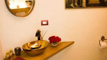 A bathroom with a round mirror, a sink with a bowl, a red towel, a trash bin, and a framed picture on the wall.