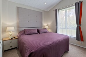 A bedroom with a large bed, a nightstand with a lamp, and a window with curtains. - St Mary's View - Luxury Lodge, Close to Beach (Tenby)
