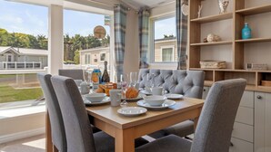 A dining area with a wooden table set for breakfast, grey upholstered chairs, and a view of a residential area through large windows. - St Mary's View - Luxury Lodge, Close to Beach (Tenby)