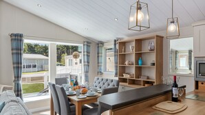 A modern kitchen with a dining area, wooden cabinets, and a large window with curtains. - St Mary's View - Luxury Lodge, Close to Beach (Tenby)