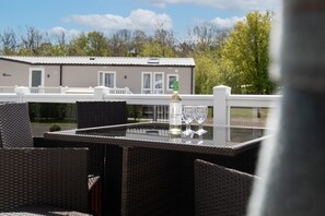 A glass-top table with wicker chairs, a bottle of wine, and two glasses on a balcony overlooking a building with a white fence and trees in the background. - St Mary's View - Luxury Lodge, Close to Beach (Tenby)