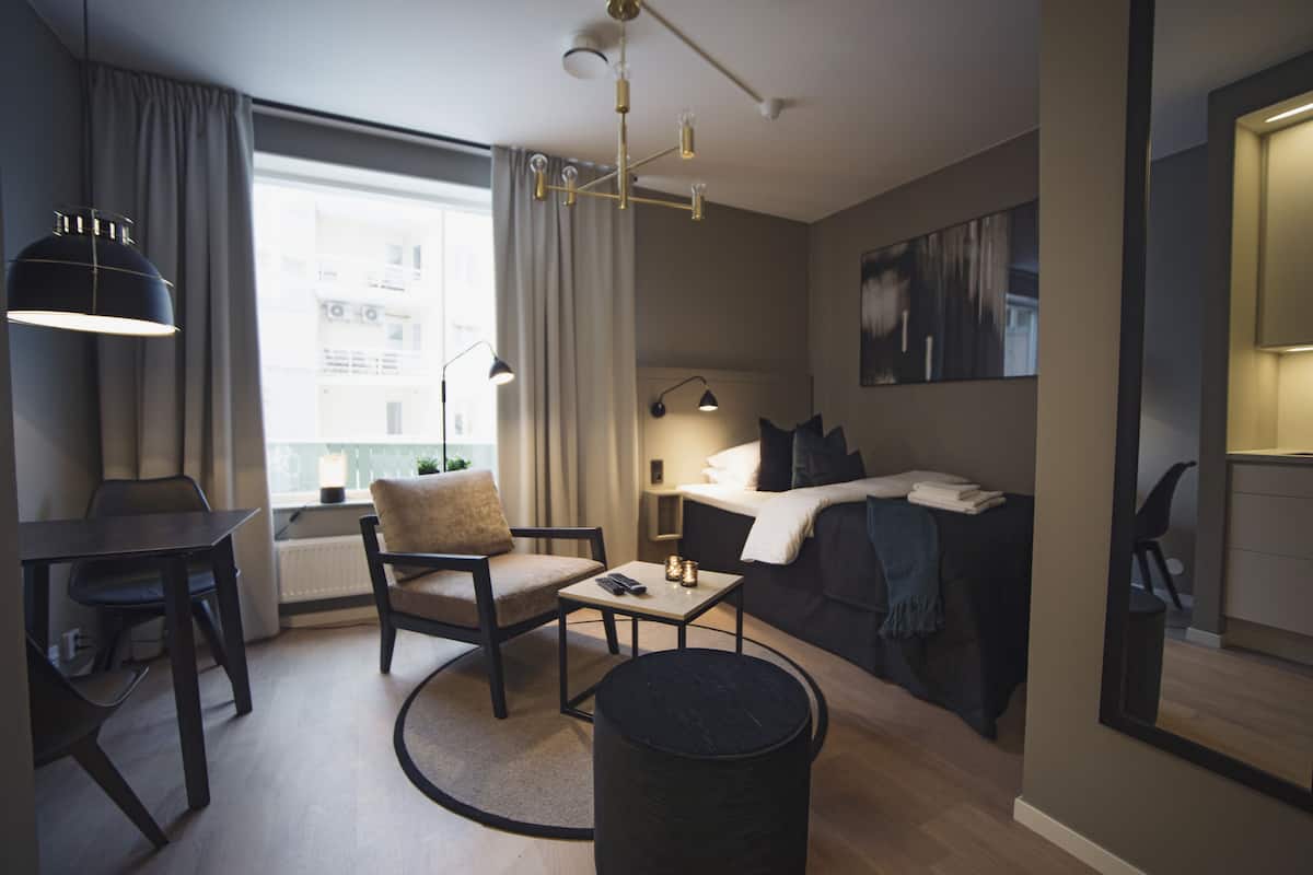 Ein modernes Hotelzimmer mit Bett, Schreibtisch, Stuhl, einem kleinen Tisch und einem runden Tisch. Ein Fenster mit Vorhängen, eine Lampe und ein Gemälde an der Wand sind ebenfalls vorhanden.