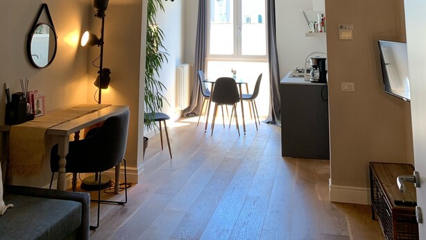 Uma sala de estar espaçosa com teto alto, uma mesa de jantar com cadeiras, um sofá, um abajur e vista através de um arco para uma janela.