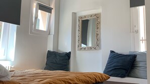 Quarto com cama, janela, espelho e um banco com sapatos.