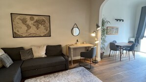 Uma sala de estar com um sofá escuro, um tapete branco, uma escrivaninha e uma mesa de jantar com cadeiras.