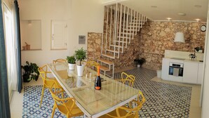 A dining area with a glass table, yellow chairs, and a patterned floor. A staircase with a wooden railing leads to a kitchen area with a stone wall. - Suite Bagnara (Syracuse)
