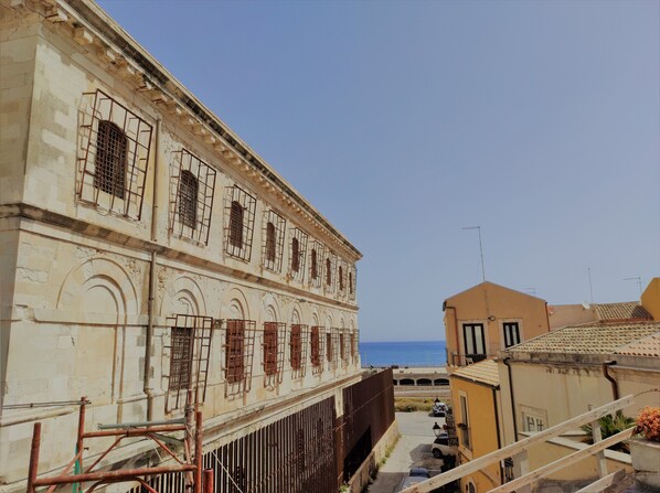 A historic building with arched windows and a balcony, overlooking a street with parked cars and a clear view of the sea.