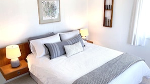 A neatly made bed with a white comforter and multiple pillows, a bedside table with a lamp, and a framed artwork on the wall.