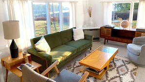 A well-lit living room with a green sofa, a wooden coffee table, and a grey armchair. There is a lamp on a side table, and a bookshelf with books and decorative items.