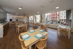 Un comedor amplio con varias mesas y sillas, ventanas grandes que ofrecen vistas a la calle y una zona de cocina con electrodomésticos modernos.