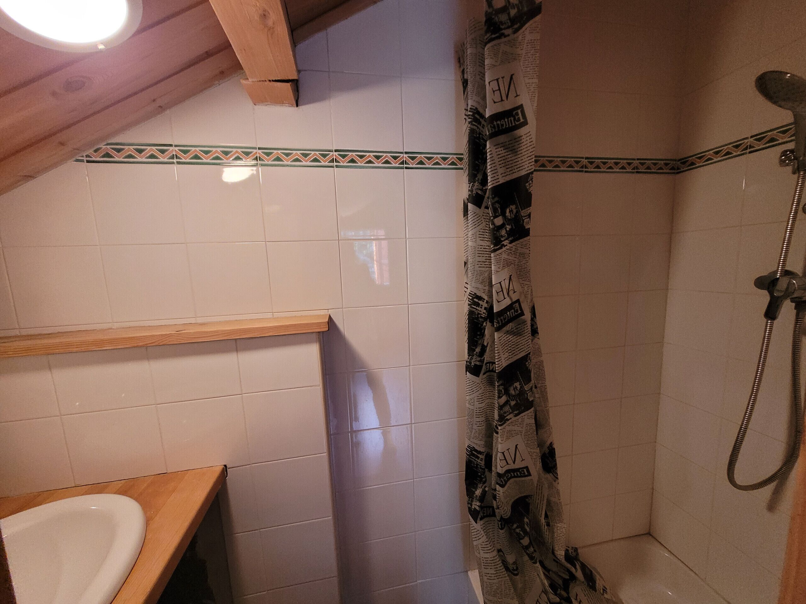 A bathroom with white tiles, a wooden shelf, a shower with newspaper curtain, and a visible ceiling light.