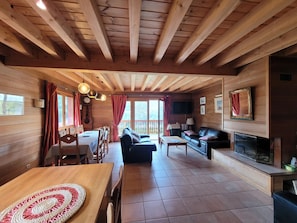 A wooden cabin interior with a dining area, sofa, fireplace, and large windows.