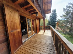 A wooden deck with a glass door leading to an interior space, a wooden railing, and a view of trees and a building in the background. - NICE 8 PERSONS SEMI-DETACHED CHALET (Saint-Chaffrey)