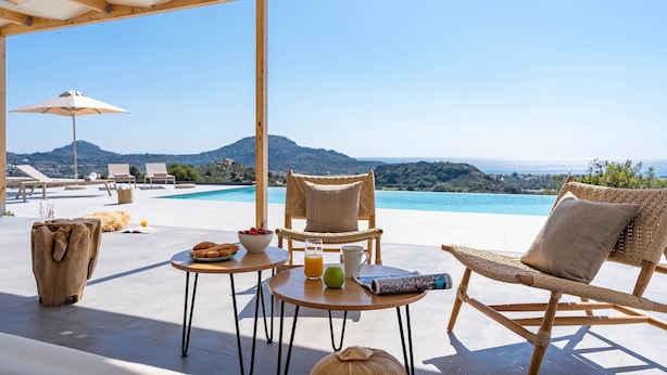 A sunlit outdoor seating area with a pool view, wicker furniture, and a breakfast setup. - Nora Villa in Afandou (Rhodes)