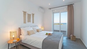 A bedroom with a bed, bedside lamps, a small table with a cup and a fruit bowl, a large window with curtains, and a view of the sea. - Nora Villa in Afandou (Rhodes)