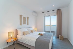 A bedroom with a bed, bedside lamps, a small table with a cup and a fruit bowl, a large window with curtains, and a view of the sea.