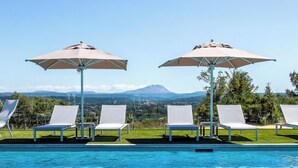 Un área de piscina con cuatro sillones blancos y dos sombrillas, rodeada por una terraza de madera.