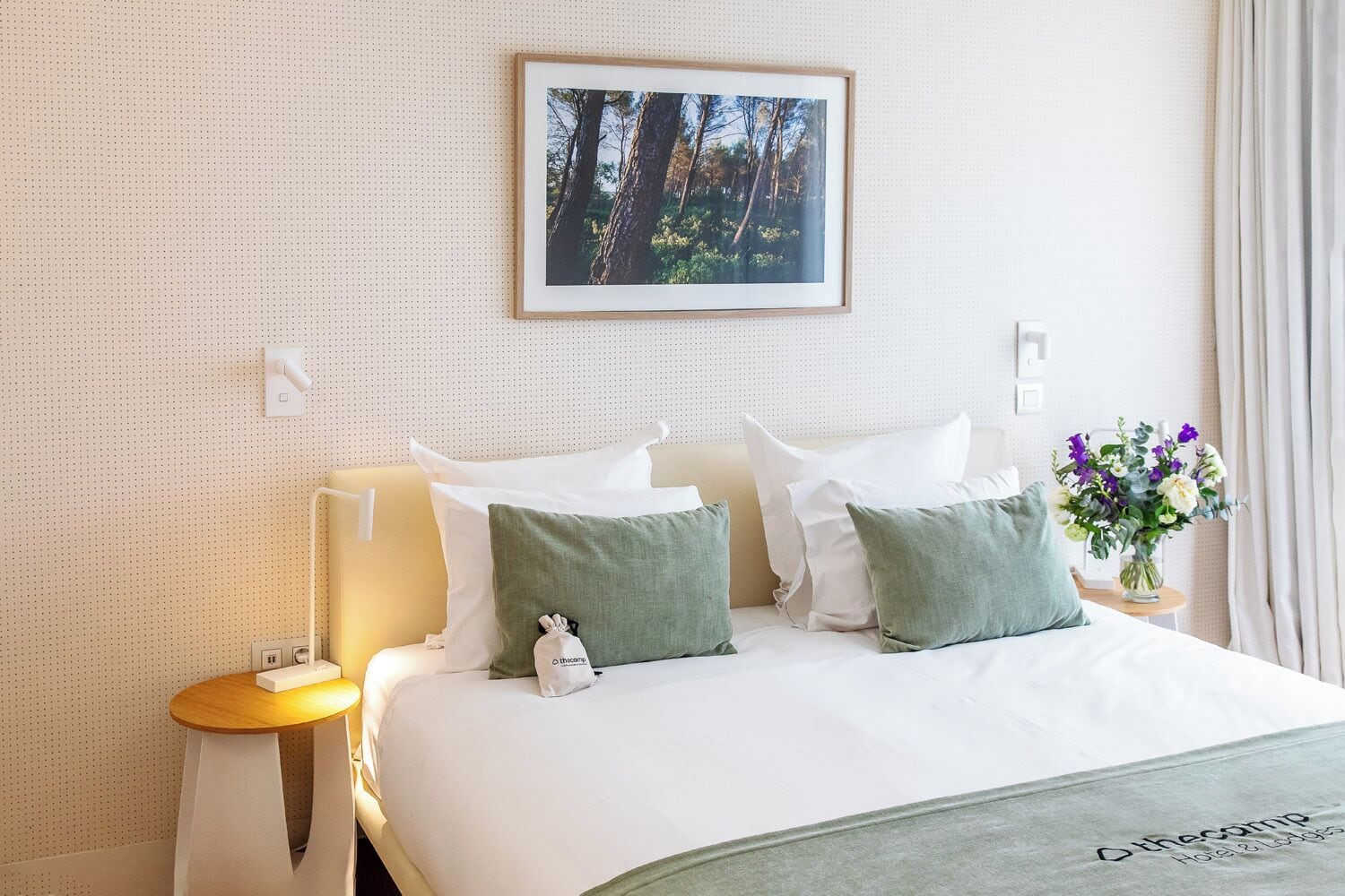 A neatly arranged hotel room with a bed, pillows, a nightstand, a framed picture on the wall, and a vase with flowers.