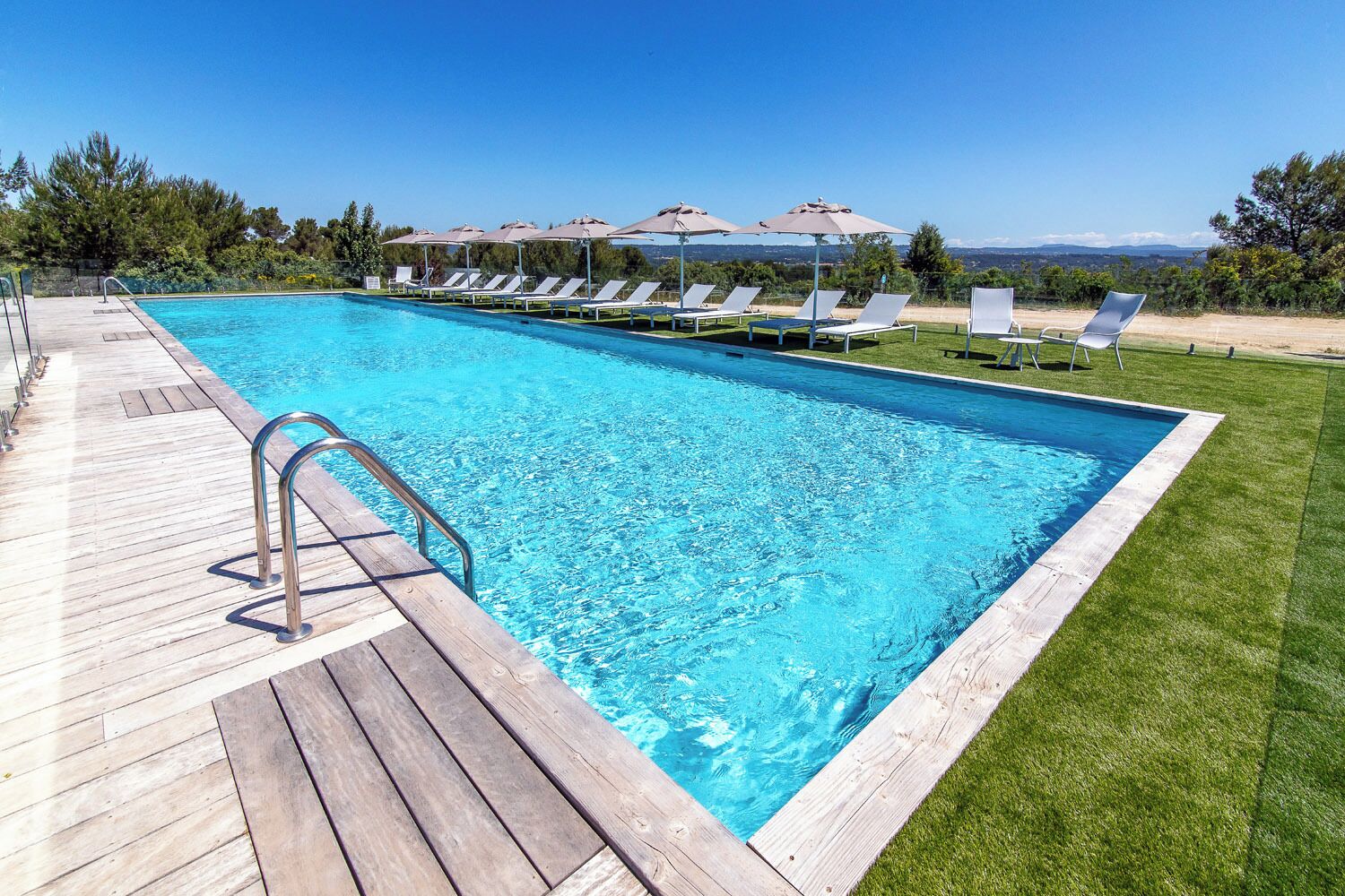 Una piscina rectangular con muelle de madera y zona de césped con sillones y sombrillas.