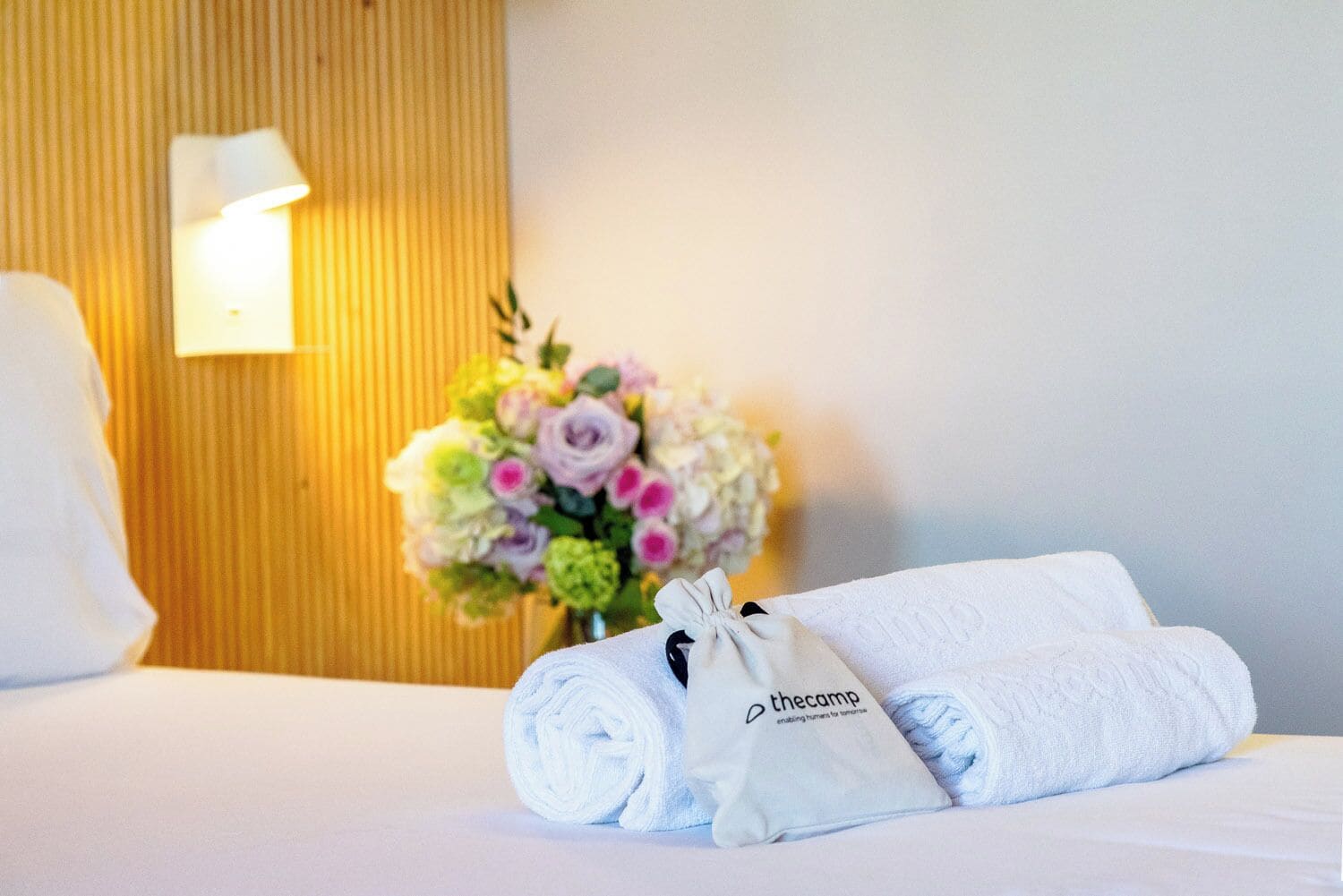 Una cama bien hecha con sábanas blancas, una toalla enrollada y una pequeña almohada blanca con un logo. Un ramo de flores está colocado sobre la cama, y se ve una lámpara fijada a la pared.