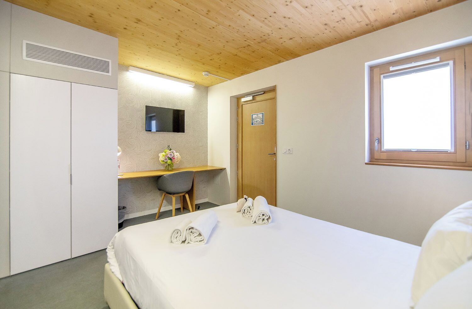 A bedroom with a bed, a desk, a chair, a TV, a wardrobe, and a window.