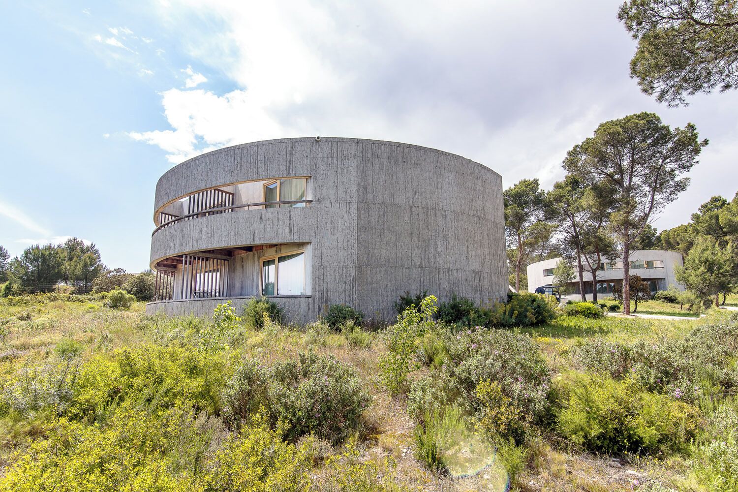 Un edificio moderno y circular con un balcón en el piso superior, rodeado de vegetación natural.